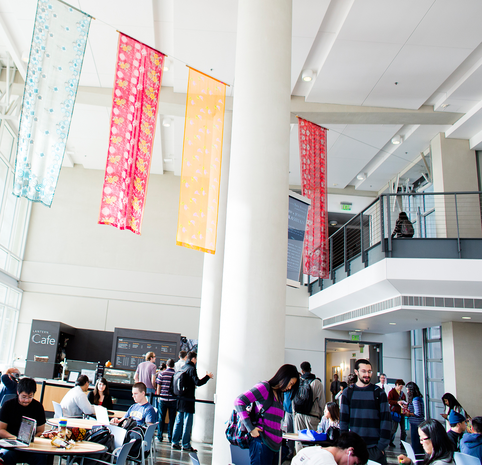A bright, modern indoor space with high ceilings and large windows shows a busy café or student common area. Several people sit at tables using laptops or talking, while others stand or walk nearby. Colorful, patterned fabric banners hang from the ceiling above. A café counter with a menu board is visible in the background, along with a sign that reads